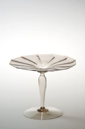  A delicate glass compote dish with a rippled edge and radial lines inside the bowl, standing on a slender stem with a round base, set against a light grey background.