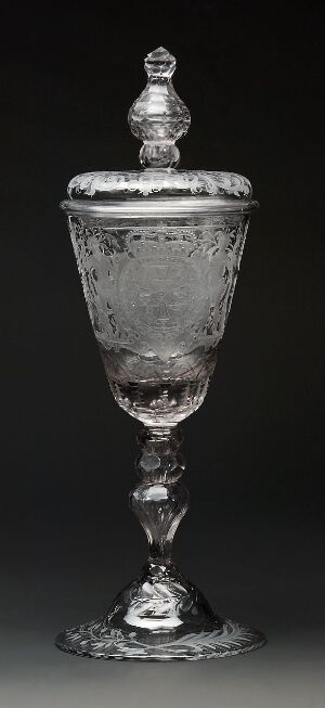  An ornate silver-colored goblet with a lid, featuring embossed or engraved decorative details on the cup and finial, exhibiting a reflective surface and standing on a dark background.