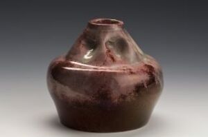  A handcrafted ceramic vessel with a bulbous body and an asymmetrical neck, glazed in deep chocolate brown with reddish-brown highlights, set against a soft, graded gray background. Artistname and title unknown.