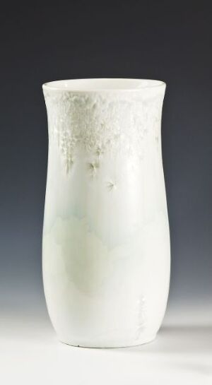  A slender, off-white ceramic vase with a subtle, speckled pattern of pale gray flecks concentrated at its top, tapering towards the base against a graduated grey background. Artist and title are unknown.