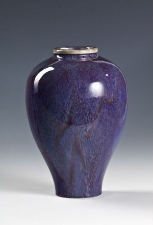  A deep blue vase with a pear-like shape and a metallic rim, set against a light grey gradient background, exhibits a subtle play of colors with areas of lighter blue and hints of purple.