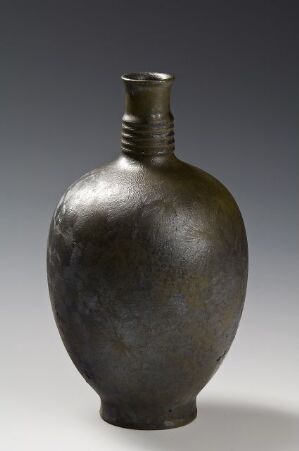  A dark gray asymmetrically shaped vase with a narrow neck and textured surface, giving it an antique metallic appearance, displayed against a light gray background. Artist name and title are unknown.