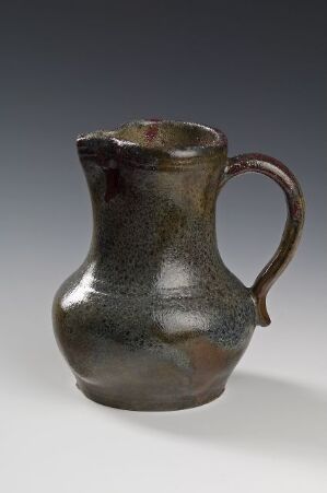  A dark-toned, glazed ceramic pitcher with a bulbous body and a curved handle, showing a shimmering blend of deep browns, bronze, and dark greens, with a slightly irregular top edge, suggesting a handmade artifact. Artist name and title are unknown.