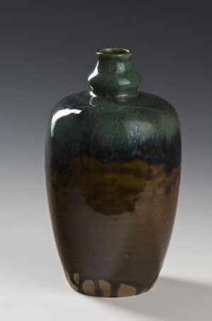  A ceramic vase with a glossy finish, featuring a dark green glaze at the top transitioning into earthy brown tones with a spotted pattern at the base, against a gradient gray background.