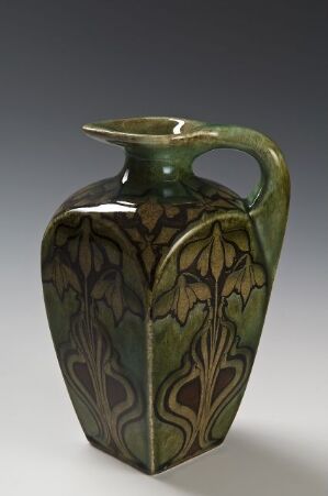  A hexagonal green art nouveau ceramic vase with a single handle and a decorative leaf design in varying shades of green, set against a muted gray background.