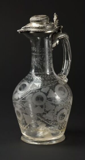  An intricately etched glass pitcher with a decorative stopper, featuring a twisted handle and floral motif designs, displayed against a black background.