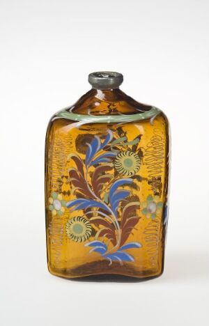  A flat, rectangular glass bottle with an ornate amber and gold floral design, featuring blue and green leaf-like patterns, against a plain, light background. Artistname and title are unknown.