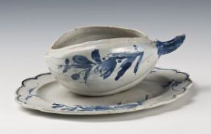  A white-glazed faience tableware set with cobalt blue hand-painted decoration by Herrebøe Fajansefabrikk, featuring an oval-shaped sauceboat with a tail-like handle poised on an accenting underplate, both adorned with matching blue flora and fauna motifs.