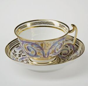  An ornate ivory teacup with golden and lavender decorations accompanied by a matching saucer, set against a plain light background.