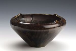  A ceramic bowl with a wavy rim, featuring a deep brown glaze with swirls of lighter brown and caramel tones against a neutral gray background, creating a lustrous and organic appearance. Artist and title are unknown.