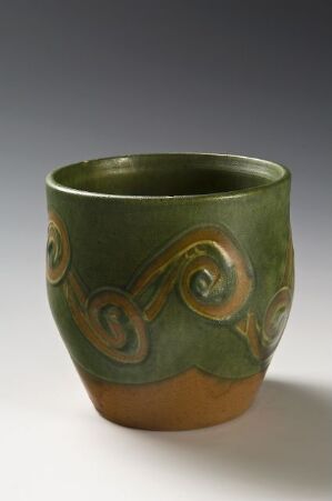  Handcrafted ceramic cup with a flared rim, featuring a deep green glaze with embossed spiral motifs on the upper portion and a raw, sandy beige base.