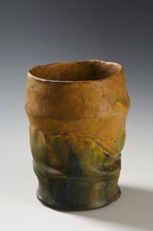 
 An earthen-toned ceramic vessel with an irregular rim stands against a soft grey background. The vessel displays a rustic glaze with transitional colors from caramel brown at the top to olive and forest greens at the base.