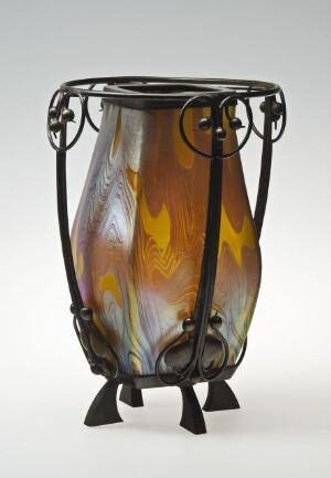  An Art Nouveau style vase made of amber to brown gradient glass with dark brown flame-like patterns and swirls of iridescent blue and green, encased in a symmetrical wrought iron frame with looping handles and a three-footed base.