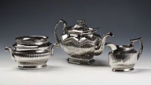  A three-piece silver tea service set with fluted design, consisting of a teapot, sugar bowl, and creamer on a neutral background.