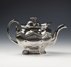  A silver teapot with embossed horizontal lines and a chevron pattern reflects light against a neutral background.