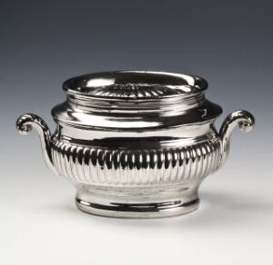  An ornate silver bowl with curved handles and horizontal ridges on the upper body, set against a neutral gray background.