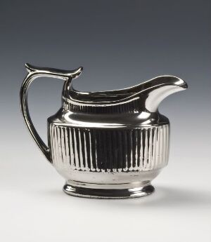  A polished silver creamer with vertical ridges, a curvilinear handle, and a graceful spout against a dark, blurred background, exuding elegance and simplicity. Artist and title are unknown.