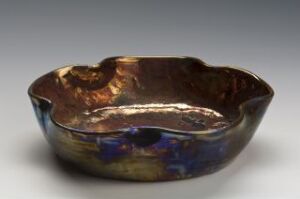  A handcrafted, asymmetrically shaped bowl with a metallic luster glaze, showcasing rich browns, blues, and purples, presented against a soft gray gradient background.