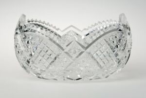  A transparent cut crystal bowl with a scalloped edge and intricate geometric patterns, reflecting light against a soft gray background. Artist name and title of the piece are unknown.