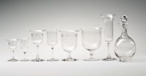  A collection of transparent glass stemware and a decanter arranged in a line against a light gray background, varying in size and shape but all clear and empty.