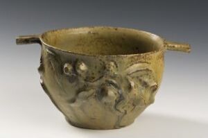  Antique ceramic bowl with raised figural designs and two handles, set against a gray background. The bowl is in an oval shape, with a beige color and darkened patches, showing textured faces in relief on its surface.