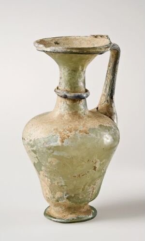  An ancient translucent green glass container titled "kanne" by an unknown artist, featuring a bulbous body with a narrowed, elongated neck and flaring mouth, and a curved handle, displaying areas of iridescence.