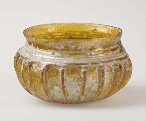  An ancient glass bowl with a flared rim and ribbed texture, displaying iridescent colors of yellow, green, purple, and blue against a light grey background. Artist name and title are unknown.