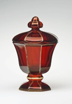  A symmetrical and geometrical lidded urn made of transparent red and amber glass, with a faceted design and a round conical foot, against a light grey background.