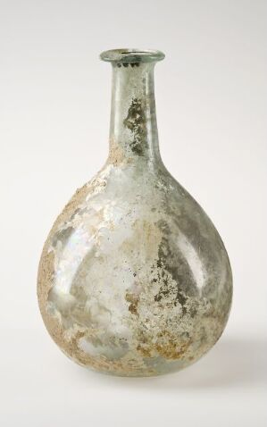  An ancient pear-shaped glass bottle with a pale green tint, showing signs of aging such as opaqueness, cloudiness, and mineral deposits against a neutral background.