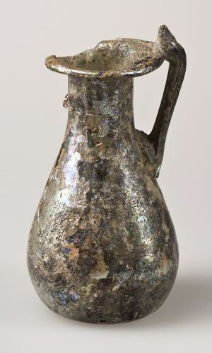  An ancient metallic pitcher with a single handle and a speckled patina of silver, gray, greenish, and brown hues, suggesting considerable age, displayed against a plain background.