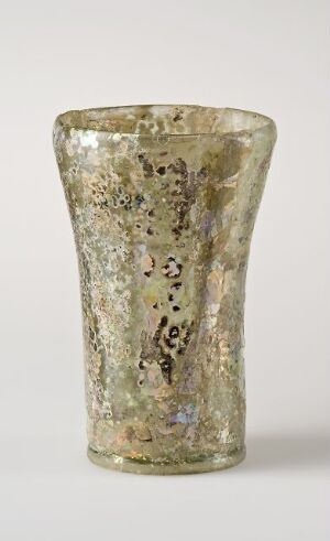  An ancient-looking glass vessel with a tapering cylindrical shape, exhibiting a silvered background and speckled with tarnish-like spots in shades of brown and black, with subtle yellow and green iridescence.