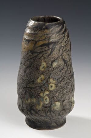  A vase with an organic, bark-like texture in dark grey with muted yellow-green splotches resembling moss, conveying an earthy and natural aesthetic. Artist name and title are unknown.