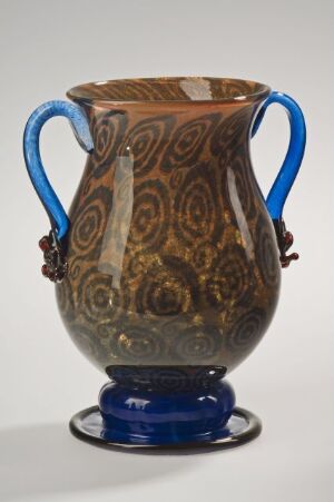  A decorative blown glass vase by Simon Gate, featuring a body with intricate peacock feather patterns in earthy tones and two prominent cobalt blue handles, resting on a coordinating blue base.