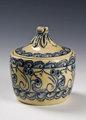  A small, round ceramic pot with a lid, featuring ornate cobalt blue floral designs on a creamy yellow background. The pot tapers slightly at the base and has intricate blue designs covering its surface, with a decorative knob atop the lid.