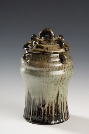 ** A handcrafted ceramic jar with a glossy brown base and mossy green top, featuring streaks of lighter brown glaze dripping down towards the darker bottom, and a lid with an ornate handle.