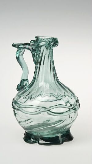  A delicate seafoam green glass pitcher with a bulbous base, elongated neck, and intricate wave pattern around the collar. The handle has an ornamental twist, and the transparent material reveals the craftsmanship of the glassblowing artistry. The pitcher is set against a gradient background transitioning from white to light grey.