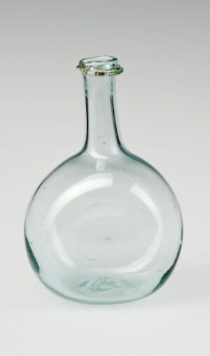  A transparent glass bottle with a pale green tint stands on a light gray background, showing a wide base, curving inward to a slender neck, and flared lip at the top. The bottle is empty and appears to be handcrafted with minor irregularities and a visible pontil mark on the base.