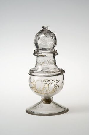  A clear glass vessel with intricate etchings on its bulbous lower body and lid, featuring a narrow neck and standing on a round base against a neutral background.