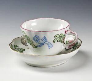  A white glazed porcelain teacup and saucer designed by Oluf Wold-Torne, featuring hand-painted overglaze decorations of a blue bird on the cup and green leaves with red rowan berries on the saucer, finished with a red rim at the edges.