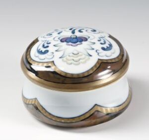  A round feldspar porcelain container adorned with cobalt blue, pale blue, and green underglaze decoration, as well as intricate gold overglaze decor on the rim and lid, designed by Hans Flygenring. The white base of the porcelain provides a stark contrast to the colorful embellishments and gold trim, creating an elegant and refined object.