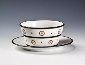  Two white feldspar porcelain bowls with black and rusty red concentric circle patterns, designed by Nora Gulbrandsen, stacked on top of each other and accompanied by a matching white plate, all set against a light grey background.