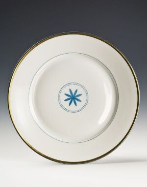  A round feldspar porcelain plate designed by Nora Gulbrandsen with a simple gold rim and a blue eight-pointed star at the center. The plate has a white background and the star provides a contrasting color element, symbolizing the piece's blend of simplicity and elegance.