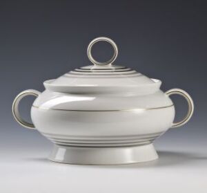  A Porsgrunds Porselænsfabrik AS feldspar porcelain soup tureen with overglaze decoration in gold and gray, featuring a bulbous body, fitted lid with circular handle, and elegant side handles, against a gray background.