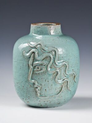  A small ceramic vase with a pale turquoise glaze and speckled texture, featuring an abstract carved design of a stylized face on its surface. The vase has a rounded body and a tapered neck, and it is displayed against a plain background.