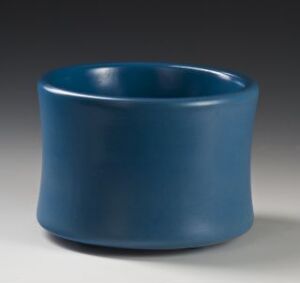  A porcelain bowl by artist Randi Gulbrandsen, featuring a glossy, cerulean blue glaze, with a cylindrical shape and an outward-flaring lip, set against a gray background emphasizing its minimalist design.