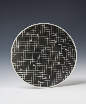  A round feldspar porcelain plate designed by Nora Gulbrandsen featuring an inscribed grid pattern with delicate star-like motifs, placed against a gradient background transitioning from light gray to black.