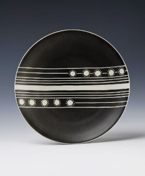  A feldspar porcelain plate designed by Nora Gulbrandsen with a glossy black surface and incised decoration. The plate features horizontal white bands with lines and evenly spaced dots, creating a simple yet elegant design.