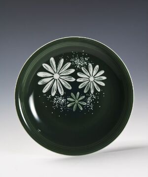  A glossy, forest green feldspar porcelain plate designed by Randi Gulbrandsen, adorned with simplistic white daisy-like flowers and scattered dot patterns in the center against a dark green background.