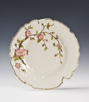  A Porsgrunds Porselænsfabrik AS porcelain plate with scalloped edges, featuring a hand-painted overglaze decoration of cherry blossoms in pink and green on a creamy white background, accented by a gold trim.