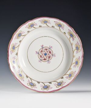  Decorative circular plate with a white background featuring a central Celtic-like knot design in shades of pink and gray, surrounded by a band of stylized blue flowers with yellow centers and soft green leaves, framed by a pink and soft purple border, displayed against a gray backdrop.